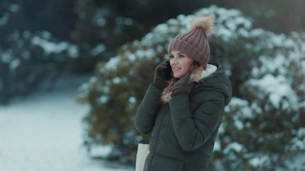 公园里穿着绿色外套 头戴褐色帽子 头戴手套 头戴便帽的快乐的现代女性在雪地的树枝边 用智能手机交谈着 — 图库视频影像