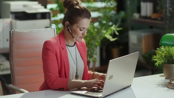 持続可能な職場 ヘッドセットとラップトップが付いている赤いジャケットの現代緑のオフィスの現代小さいビジネスオーナーの女性を微笑みます — ストック動画