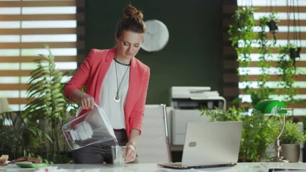 持続可能な職場 水フィルター ラップトップのカップと赤いジャケットで仕事をしている現代の小さなビジネスオーナーの女性を微笑みます — ストック動画