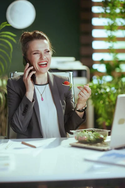 Sustainable workplace. smiling modern accountant woman in a grey business suit in modern green office with laptop eating salad and talking on a smartphone.