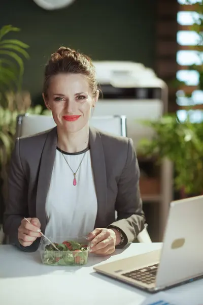 Sustainable workplace. modern 40 years old small business owner woman in a grey business suit in modern green office with laptop eating salad.