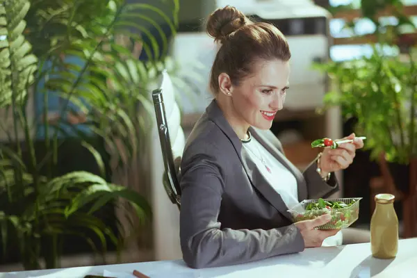 Sustainable workplace. happy modern middle aged woman worker in a grey business suit in modern green office eating salad.