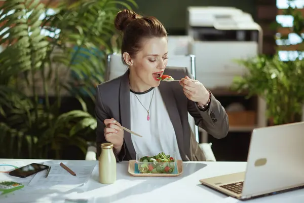 Sustainable workplace. modern small business owner woman in a grey business suit in modern green office with laptop eating salad.