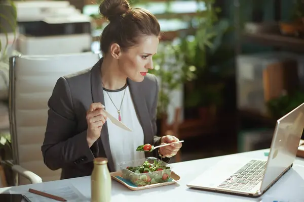可持续工作场所 穿着灰色西装 身穿现代绿色办公室工作服 手持笔记本电脑 吃沙拉的中年妇女 — 图库照片