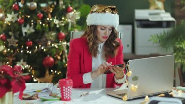 Noel zamanı. Gülümseyen, Noel Baba şapkalı, kırmızı ceketli, akıllı telefon uygulaması kullanan ve modern yeşil ofiste Noel ağacıyla çevrimiçi toplantı yapan modern, küçük işletme sahibi kadın..
