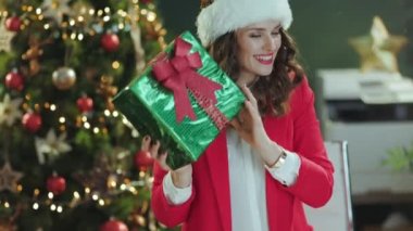 Noel zamanı. Gülümseyen 40 yaşındaki zarif iş kadını Noel Baba şapkası ve kırmızı ceketli modern yeşil ofisindeki hediye kutusuyla Noel ağacı olan..