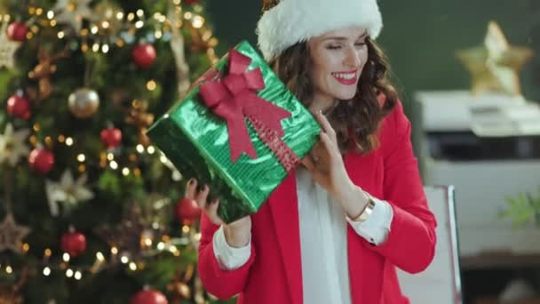 クリスマス タイム エレガントな ビジネス女性 サンタ帽子と赤いジャケット クリスマスツリーと現代緑のオフィスの現在のボックス — ストック動画