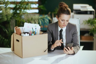 Yeni bir iş. Gri takım elbiseli, mukavva kutusunda kişisel eşyaları olan, modern yeşil ofisteki 40 yıllık modern kadın çalışan akıllı telefondan konuşuyor..