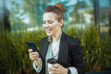 Gülümseyen, 40 yaşında, ofis yakınında çalışan, siyah ceketli, akıllı telefon kullanan, bir fincan kahveli bir kadın..