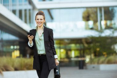 smiling modern 40 years old woman employee near business center in black jacket with smartphone and briefcase walking. clipart