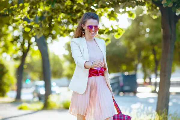 城市里快乐的中年妇女 身穿粉色衣服 身穿白色夹克 戴着太阳镜走路 — 图库照片