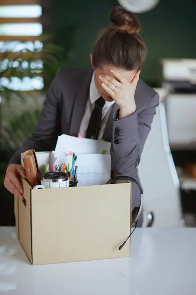 New job. sad modern female employee in modern green office in grey business suit with personal belongings in cardboard box.