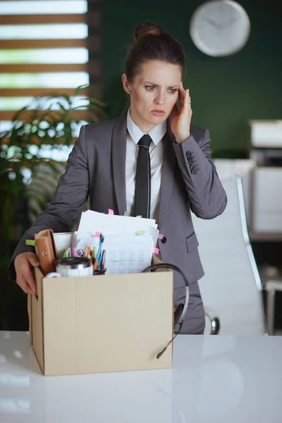 stock image New job. pensive modern woman worker in modern green office in grey business suit with personal belongings in cardboard box.