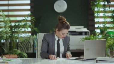 Yeni bir iş. Modern yeşil ofisteki, gri takım elbiseli, modern orta yaşlı kadın çalışanın dizüstü bilgisayarına dökümanlar fırlatıyor..