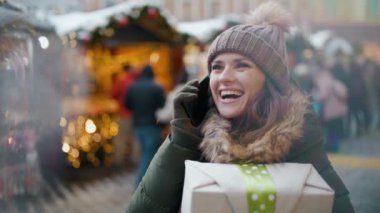 Şehirdeki kış fuarında yeşil ceketli ve kahverengi şapkalı gülümseyen genç bir bayan akıllı telefondan Noel hediyesiyle konuşuyor..