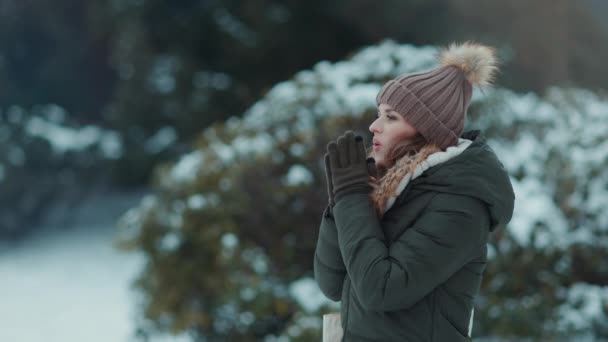 緑のコートと冬の街公園の屋外の茶色の帽子とビーニーは息で冷たい手を暖めます — ストック動画