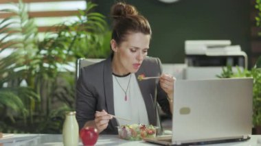 Sürdürülebilir iş yeri. Modern yeşil ofiste gri takım elbiseli, orta yaşlı bir muhasebeci kadın dizüstü bilgisayarla salata yiyor..