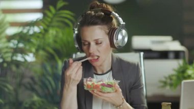 Sürdürülebilir iş yeri. Modern yeşil ofisteki gri takım elbiseli, modern 40 yaşındaki gülümseyen muhasebeci kadın. Salata yiyor ve kulaklıkla müzik dinliyor..