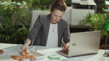 Sürdürülebilir iş yeri. Modern yeşil ofiste, gri takım elbiseli, pizza ve dizüstü bilgisayarlı modern orta yaşlı kadın işçi..