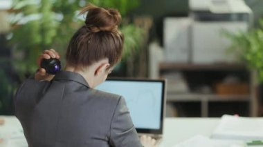 Sürdürülebilir iş yeri. Gri takım elbiseli, modern yeşil ofisteki modern iş kadınının arkasında boynundaki masaj silindirini kullanırken görülüyor..