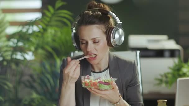 持続可能な職場 現代の笑顔40歳のブックキーパーの女性ノートパソコンでサラダを食べヘッドフォンで音楽を聴く現代の緑のオフィスでグレーのビジネススーツ — ストック動画