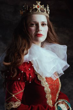 Kırmızı elbiseli, beyaz yakalı ve tacı koyu gri arka planlı ortaçağ kraliçesi..