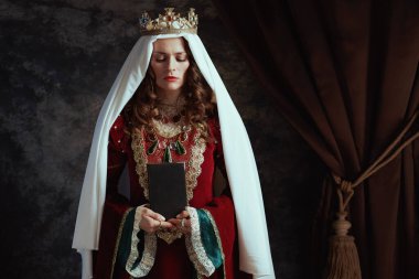 Kırmızı elbiseli, kitap, peçe ve tacı koyu gri arka planda olan ortaçağ kraliçesi.