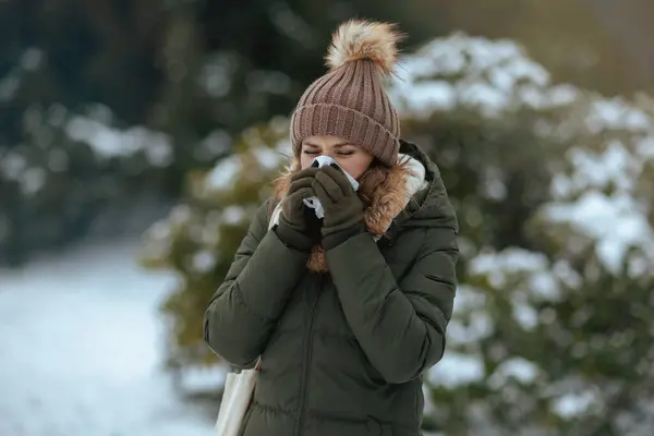 unhappy modern woman in green coat and brown hat outdoors in the city park in winter with mittens, napkin blowing nose and beanie hat.