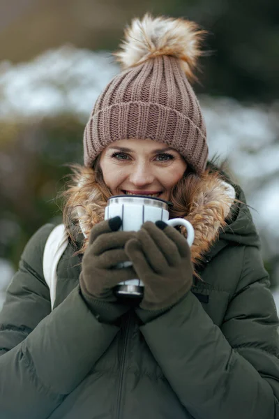 smiling modern 40 years old woman in green coat and brown hat outdoors in the city park in winter with mittens, cup of tea and beanie hat.