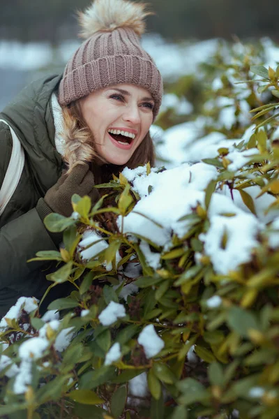 快乐的中年妇女冬天穿着绿色外套 戴着棕色帽子 头戴便帽 头戴雪白的枝头 站在城市公园的室外 — 图库照片