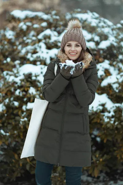 冬日里 穿着绿色外套 戴着棕色帽子 头戴燕尾服 头戴燕尾服的现代女性在城市公园的雪地枝头边微笑着 — 图库照片