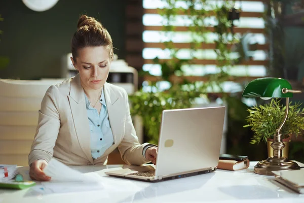 modern middle aged accountant woman in a light business suit in modern green office with documents and laptop.
