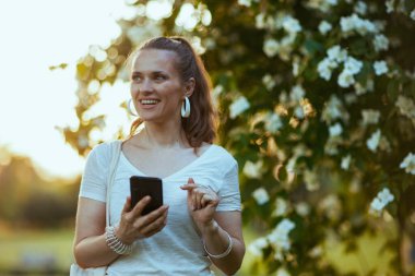 Yaz zamanı. Çiçek açan ağacın yanında akıllı telefon uygulamaları kullanarak beyaz gömlekli genç bir bayan..