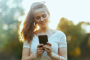 Yaz zamanı. Beyaz tişörtlü zarif bir bayan şehir parkında akıllı telefon uygulaması kullanıyor..