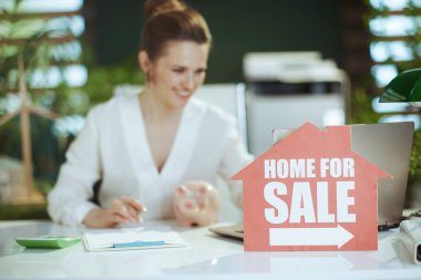 Sürdürülebilir emlak işi. gülümseyen modern kadın emlakçı beyaz bluz içinde modern yeşil ofis içinde ev tabelası, domuz kumbarası, hesap makinesi ve belge.