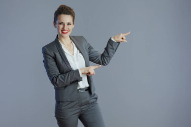 Gri takım elbiseli mutlu, orta yaşlı kadın çalışanı gri arka plandaki bir şeyi işaret ediyor..