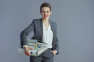 Gri arkaplanda klasörleri olan gri takım elbiseli mutlu stil sahibi bir kadın işçi..