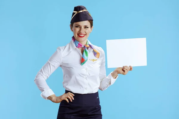 happy modern flight attendant woman isolated on blue background in uniform showing blank a4 paper sheet.