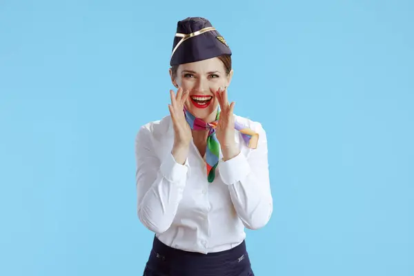 happy modern flight attendant woman on blue background in uniform shouting through megaphone shaped hands.