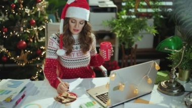 Noel zamanı. Noel Baba şapkalı, kırmızı Noel süveterli, geleneksel kurabiyeli, modern yeşil ofiste şarkı söyleyen, güler yüzlü, şık, küçük işletme sahibi kadın..