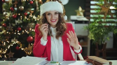 Noel zamanı. Kırmızı ceketli gülümseyen, video konuşan, modern yeşil ofiste Noel ağacıyla Noel Baba şapkasıyla konuşan..