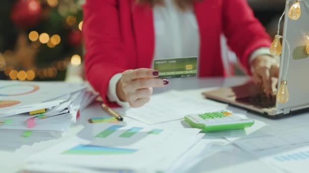 クリスマス タイム クリスマスツリーでグリーンオフィスで働く文書 ラップトップ クレジットカード付きのサンタ帽子と赤いジャケットのビジネス女性のクローズアップ — ストック動画