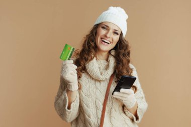 Merhaba kış. Bej kazaklı, eldivenli ve şapkalı gülümseyen zarif bir kadının portresi. Akıllı telefon ve kredi kartı takılı..