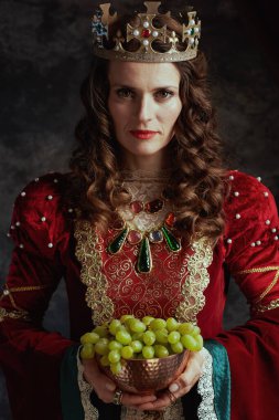 Kırmızı elbiseli ortaçağ kraliçesi. Üzüm tabağı ve tacı koyu gri arka planda..