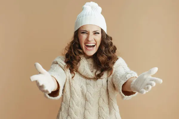 Hello winter. smiling stylish middle aged woman in beige sweater, mittens and hat welcoming isolated on beige.