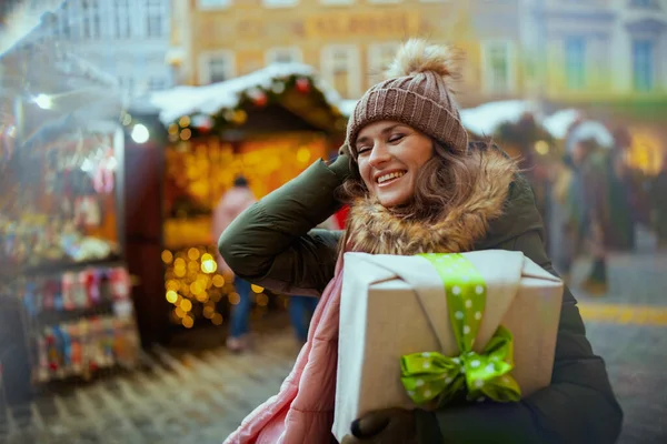 happy young woman in green coat and brown hat at the christmas fair in the city with christmas gift.