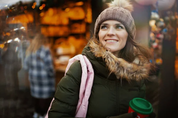smiling modern woman in green coat and brown hat at the winter fair in the city with red cup of hot Christmas beverage.