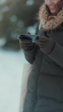 Kışın şehir parkında, akıllı telefon uygulaması kullanan eldivenli mutlu bir kadına yakın çekim..