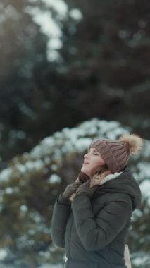 Gülümseyen, yeşil ceketli, kahverengi şapkalı modern bir kadın kışın şehir parkında eldivenli ve bereli..