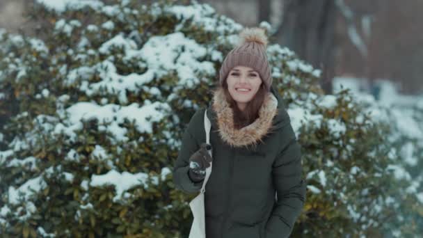 緑のコートで幸せな現代中年女性と冬の街公園の屋外でミトンと雪の枝の近くのビーニー帽子 — ストック動画